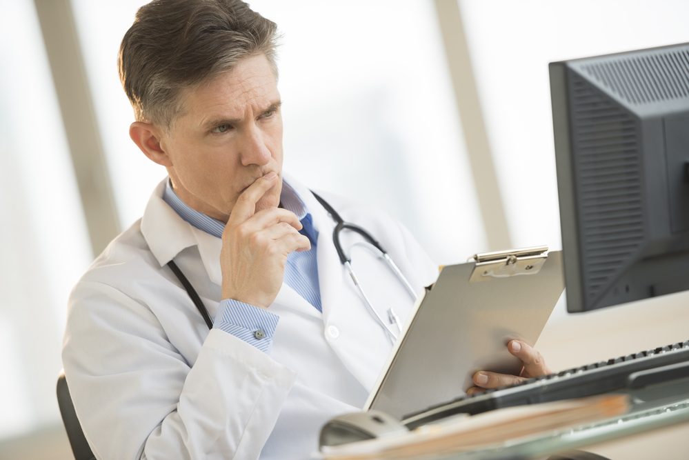 Serious mature male doctor looking at computer monitor while holding clipboard at desk in clinic